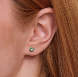 Model wears dainty flower shape diamond earring stud with green emeralds in solid 14k gold. 